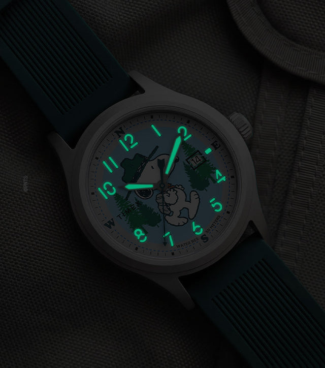 Peanuts® x Timex Expedition® Scout watch with illuminated green Indiglo® dial, highlighting the glowing Arabic numerals, hour markers, and hands against a dark gray tactical fabric background.