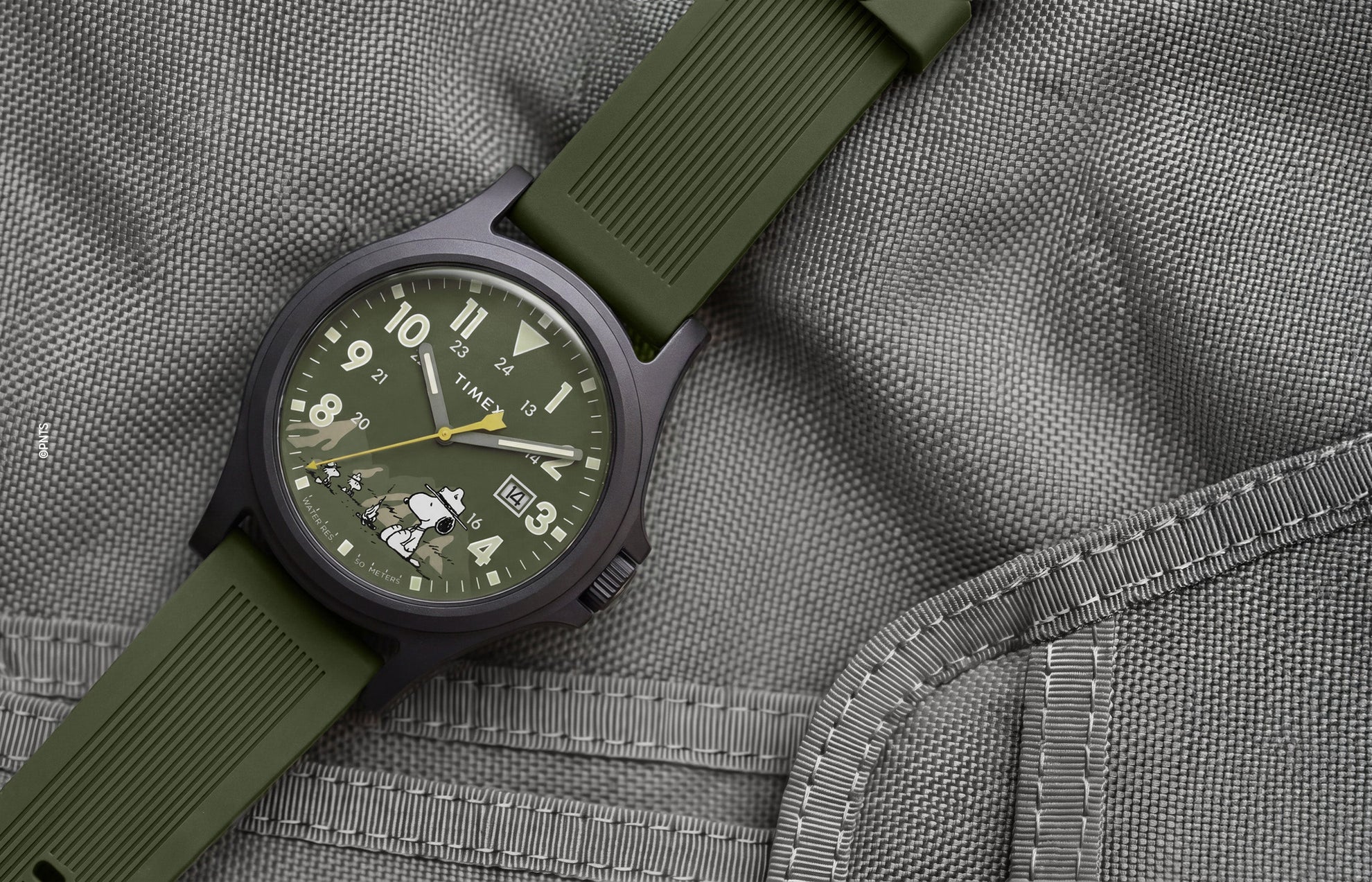 Peanuts® x Timex Expedition® Acadia watch with a matte black case, olive green dial and silicone strap, featuring Snoopy in camouflage artwork, bold Arabic numerals, a date window, and yellow seconds hand displayed on a gray tactical fabric background.