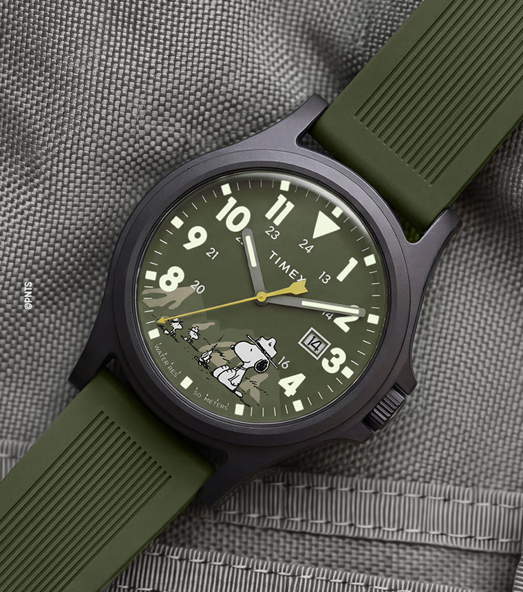 Peanuts® x Timex Expedition® Acadia watch with a matte black case, olive green dial and silicone strap, featuring Snoopy in camouflage artwork, bold Arabic numerals, a date window, and yellow seconds hand displayed on a gray tactical fabric background.