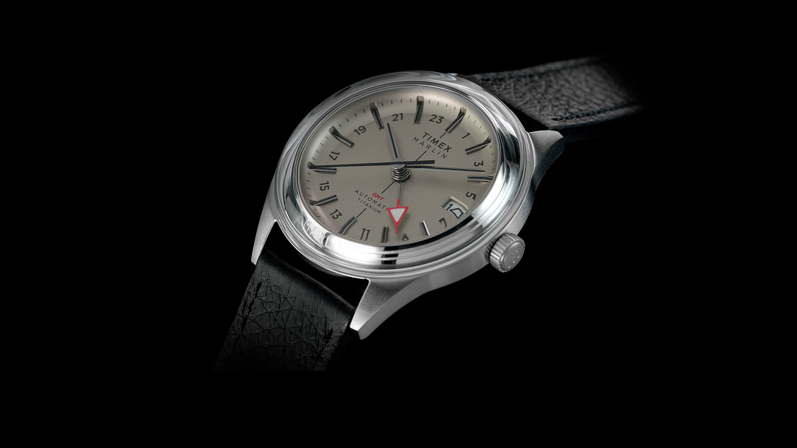Angled view of the Timex Marlin® Jet Titanium Automatic GMT featuring a brushed titanium case, cream dial, black leather strap, and red GMT hand.