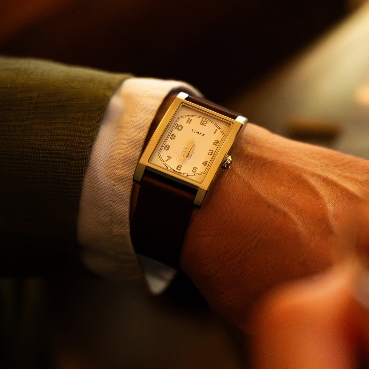 TWG069000 Close-up of a WM Brown x Timex watch with a rectangular case and brown leather strap, worn beside a shirt cuff in warm light.