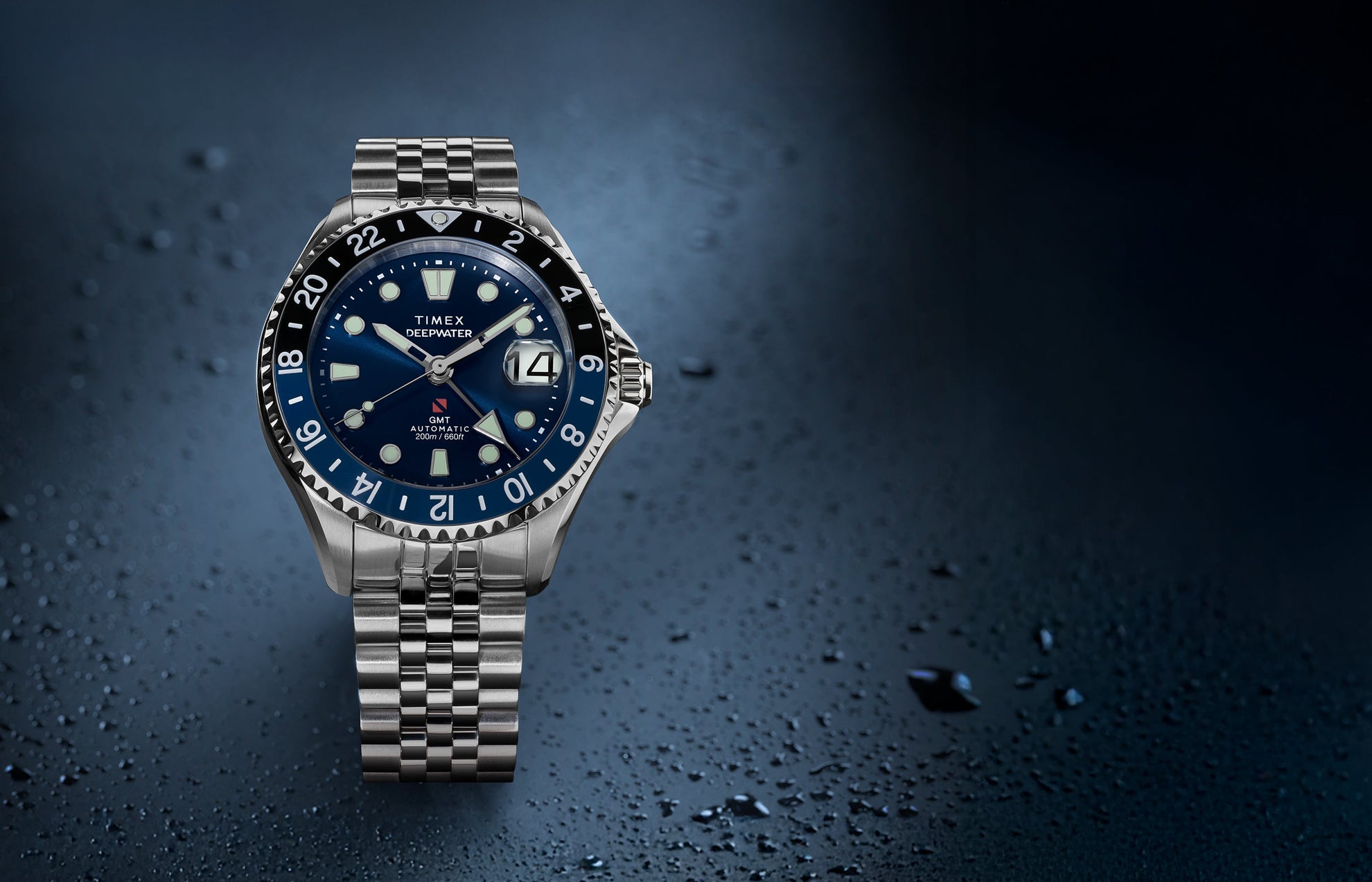 Timex Deepwater Meridian 200 Automatic GMT stainless steel watch featuring a blue sunray dial, GMT bezel, magnified date window, and jubilee-style bracelet on a textured blue background.