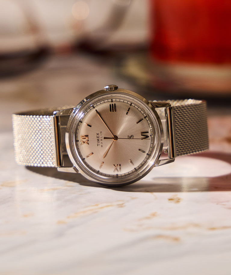 Todd Snyder x Timex Marlin® Mesh watch with a silver sunray dial, polished stainless steel case, Roman numeral hour markers, and stainless steel mesh bracelet, displayed on a marble surface with a softly blurred warm-toned background.