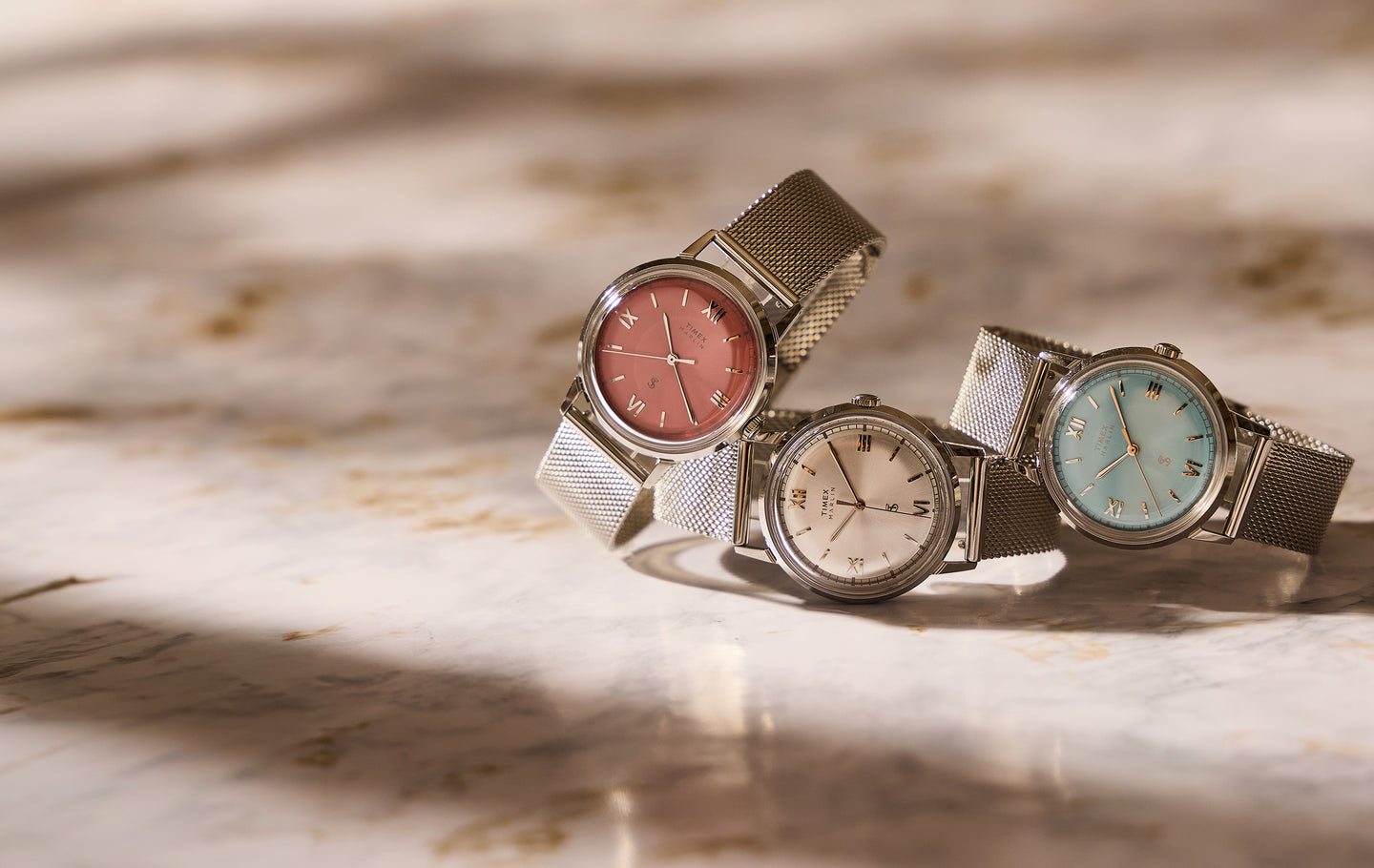 Todd Snyder x Timex Marlin® Mesh watches with pink, silver, and light blue dials and stainless steel mesh bracelets displayed together on a marble surface.