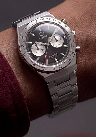 Q Timex Continental Chronograph worn on the wrist with a burgundy jacket, featuring a black chronograph dial and stainless-steel bracelet.