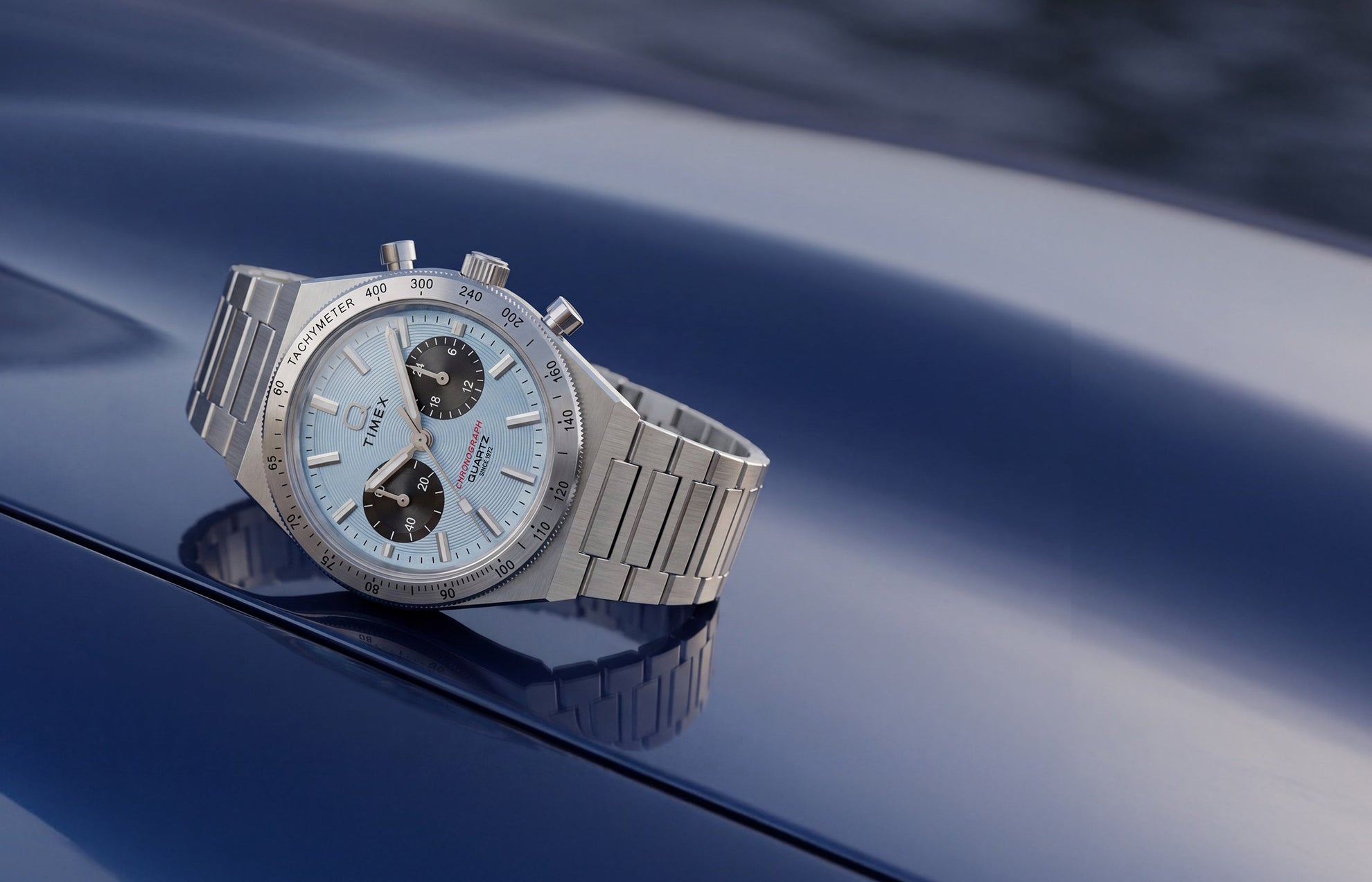 Q Timex Continental Chronograph with an ice blue dial, black subdials, and stainless-steel bracelet resting on a reflective blue surface.