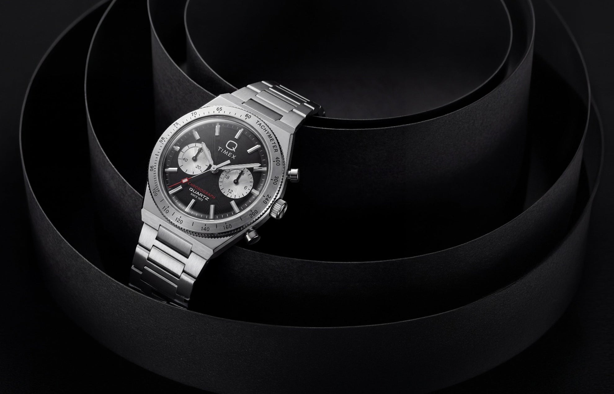 Q Timex Continental Chronograph with a black dial, silver-tone subdials, and stainless-steel bracelet displayed against sculptural black curves.
