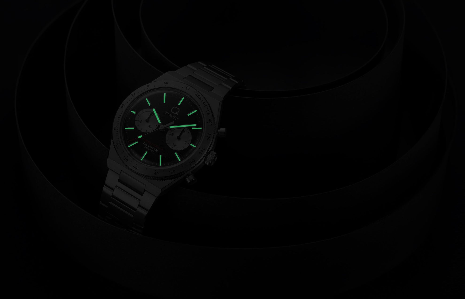 Q Timex Continental Chronograph in low light with green luminous hands and hour markers glowing against a black background.