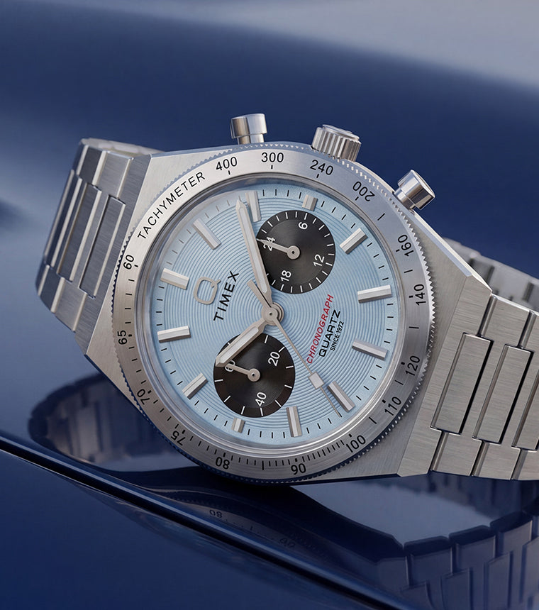 Q Timex Continental Chronograph with an ice blue dial, black subdials, and stainless-steel bracelet resting on a reflective blue surface.