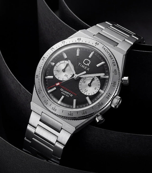 Q Timex Continental Chronograph with a black dial, silver-tone subdials, and stainless-steel bracelet displayed against sculptural black curves.