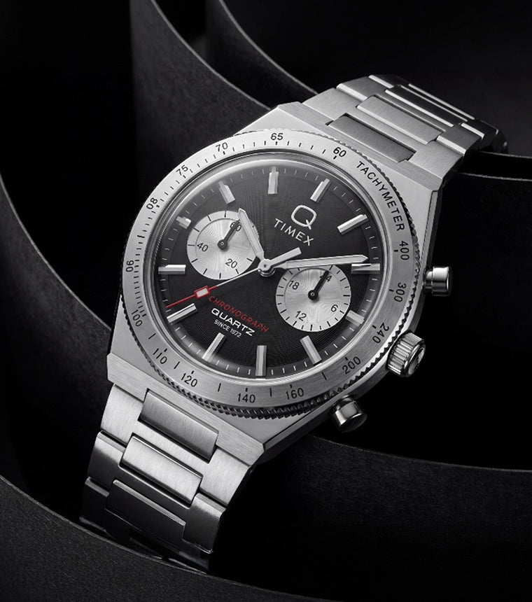 Q Timex Continental Chronograph with a black dial, silver-tone subdials, and stainless-steel bracelet displayed against sculptural black curves.
