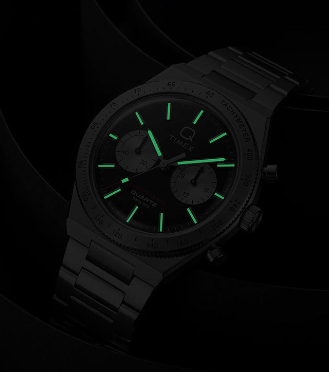 Q Timex Continental Chronograph in low light with green luminous hands and hour markers glowing against a black background.