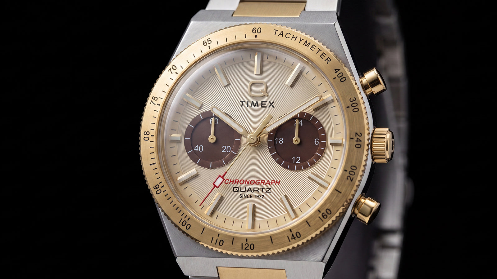 Close-up of the Q Timex Continental Chronograph featuring a champagne dial, brown subdials, gold-tone tachymeter bezel, and red chronograph hand.