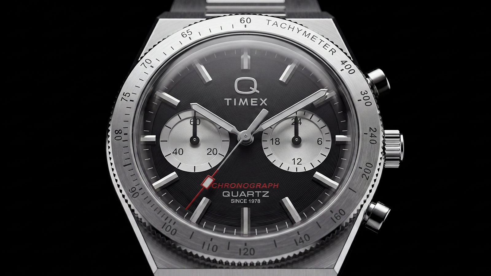 Close-up of the Q Timex Continental Chronograph featuring a black textured dial, silver-tone subdials, red chronograph hand, and stainless-steel tachymeter bezel.