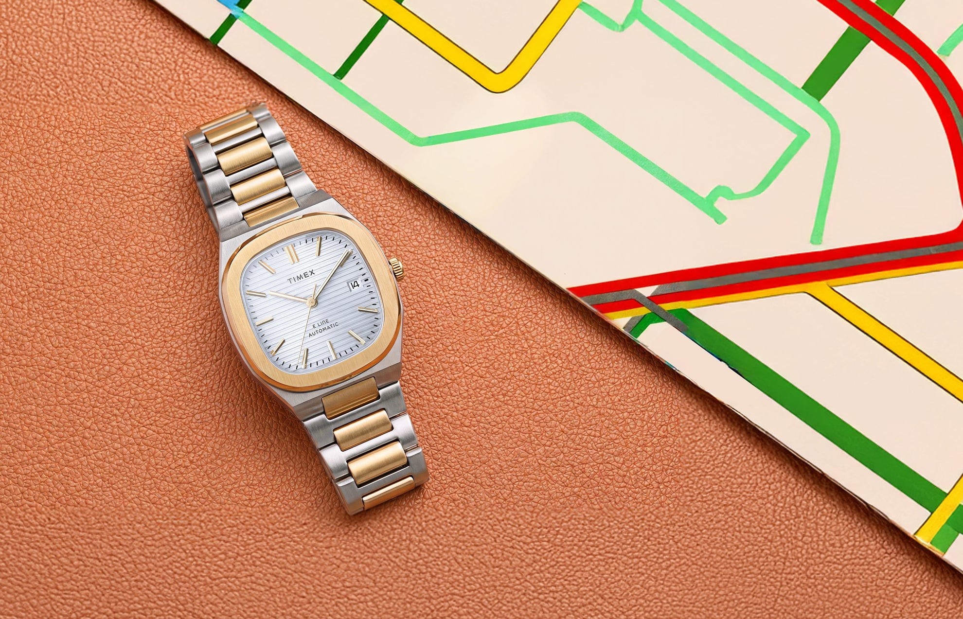Timex® Automatic E Line 38mm watch with a two-tone stainless steel bracelet, gold-tone bezel, silver textured dial, and date window displayed beside a colorful transit map.