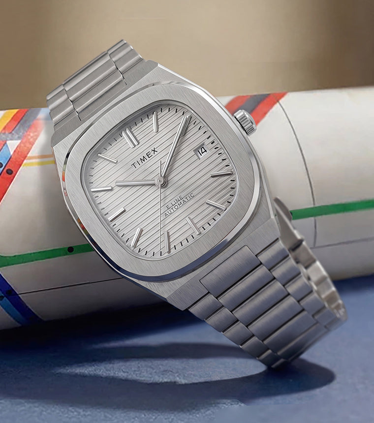Timex® Automatic E Line 38mm watch with a stainless steel bracelet, silver textured dial, and date window displayed on a rolled New York City subway map.