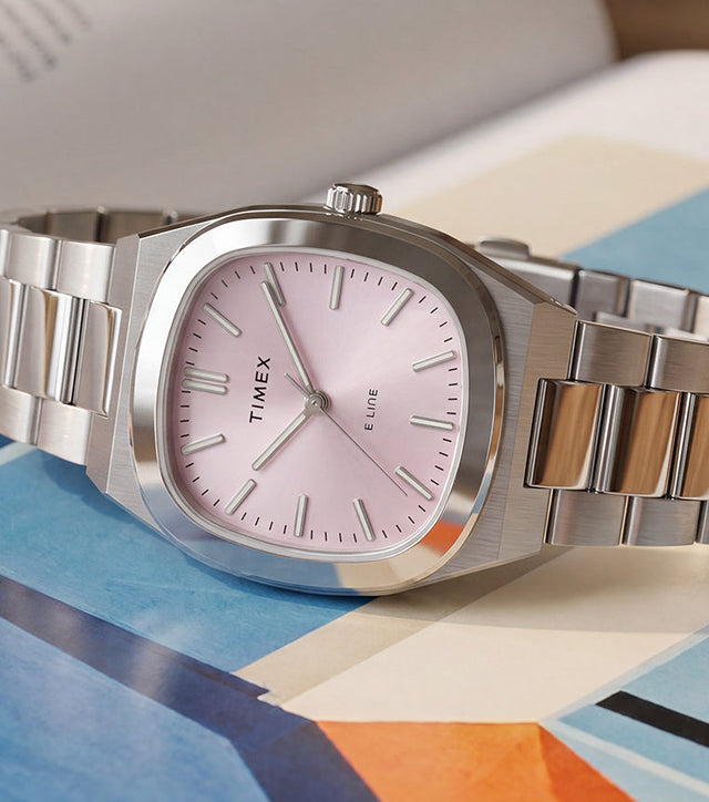 Timex® Quartz E Line 30mm watch with a stainless steel bracelet and pink sunray dial displayed on an open book with colorful artwork.