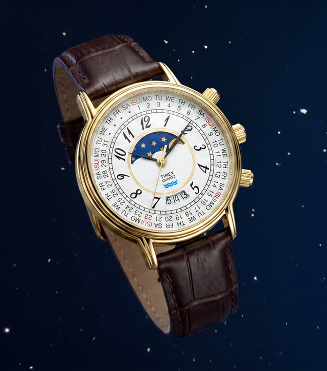 Timex 1990 Moon Phase and Calendar Dial Reissue with a gold-tone case, white calendar dial, moon phase display, and brown leather strap against a starry blue background.