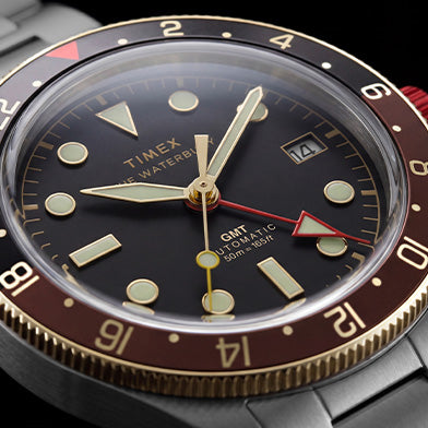 Close-up of the Waterbury Heritage Automatic GMT watch face, featuring a black dial, brown bezel, red GMT hand, and date window.