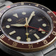 Close-up of the Waterbury Heritage Automatic GMT watch face, featuring a black dial, brown bezel, red GMT hand, and date window.