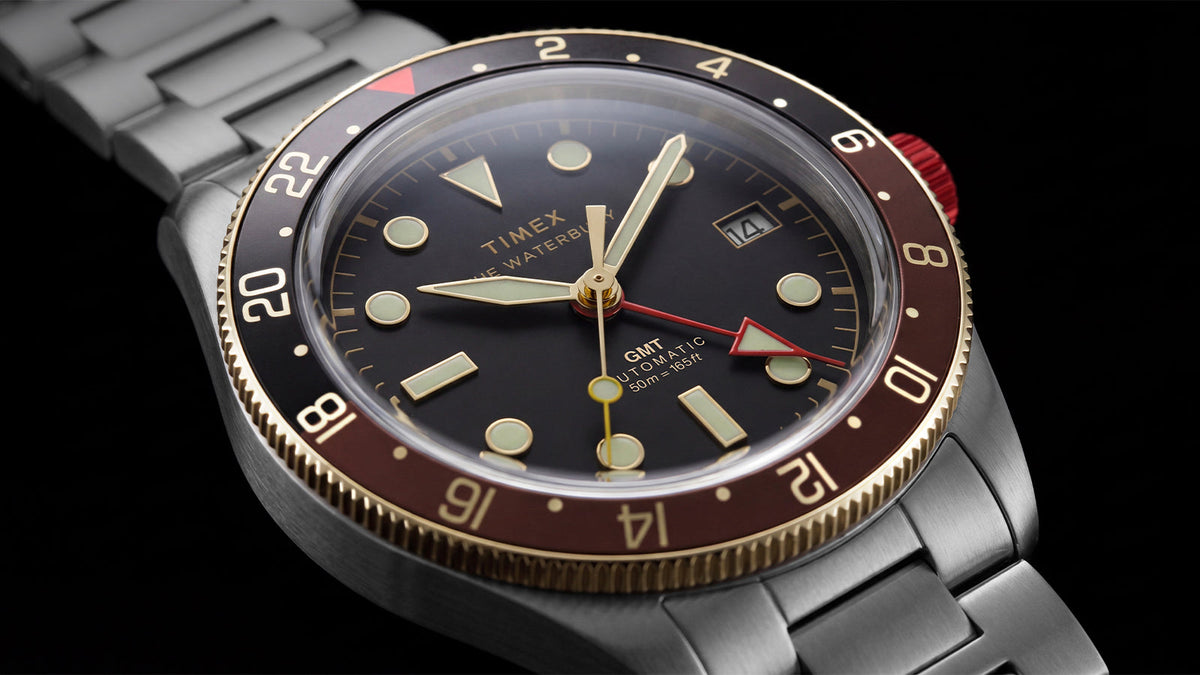 Angled close-up of the Waterbury Heritage Automatic GMT watch face and stainless-steel bracelet against a black background.