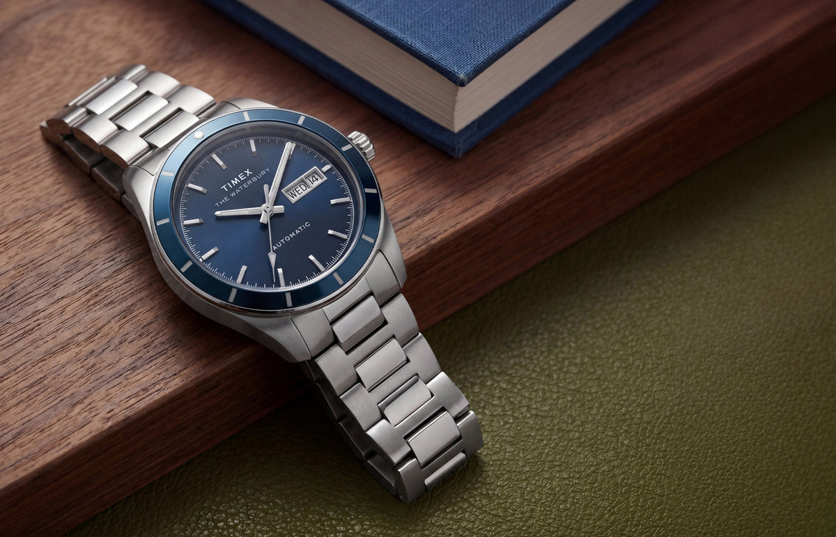 Waterbury Heritage Automatic watch with a blue dial and stainless-steel bracelet resting beside a blue book on a wooden surface.