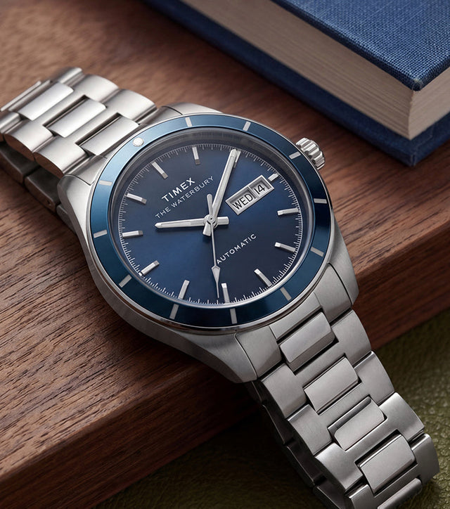 Waterbury Heritage Automatic watch with a blue dial and stainless-steel bracelet resting beside a blue book on a wooden surface.