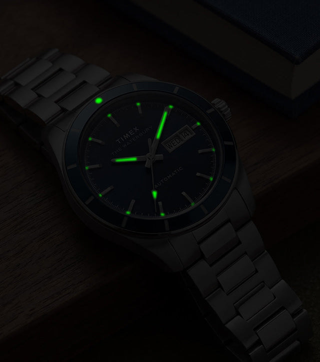 Waterbury Heritage Automatic watch glowing green in low light beside a blue book on a wooden surface.