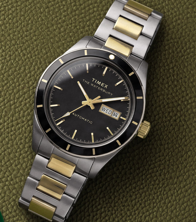 Waterbury Heritage Automatic watch with a black dial and two-tone bracelet resting on textured olive-green leather beside a golf tag.