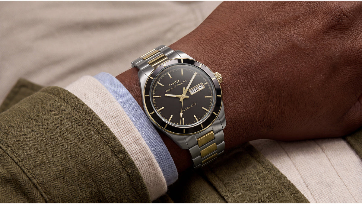 Waterbury Heritage Automatic watch with a black dial and two-tone stainless-steel bracelet worn with an olive-green jacket.
