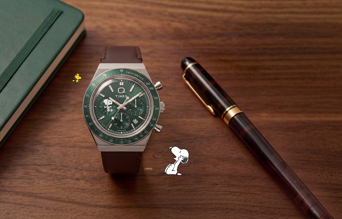 Peanuts® x Q Timex Chronograph with a green dial, Snoopy subdial, and brown leather strap displayed beside a notebook and pen.