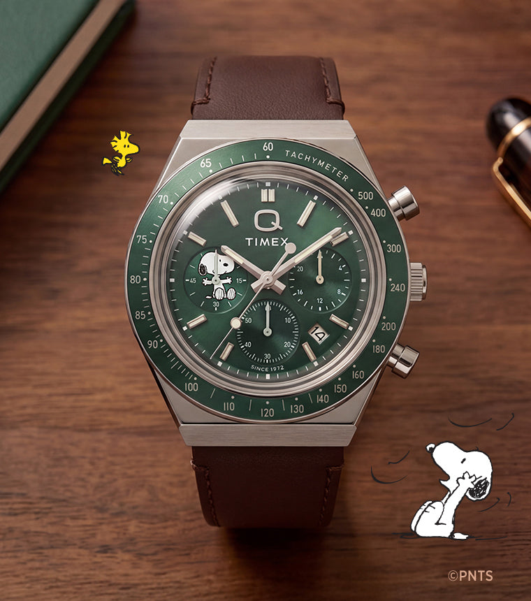 Peanuts® x Q Timex Chronograph with a green dial, Snoopy subdial, and brown leather strap displayed beside a notebook and pen.