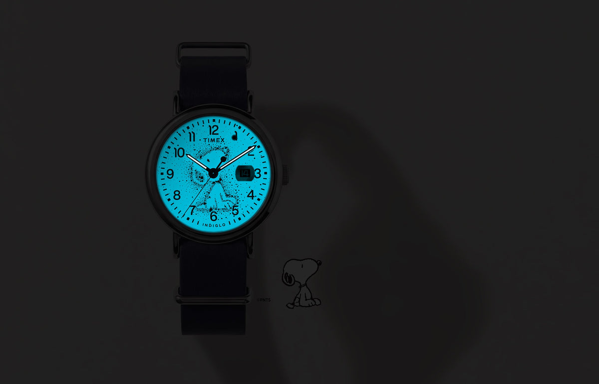 Peanuts® x Timex Weekender Astronomy watch shown in the dark with its blue INDIGLO® dial illuminating the Snoopy constellation design.