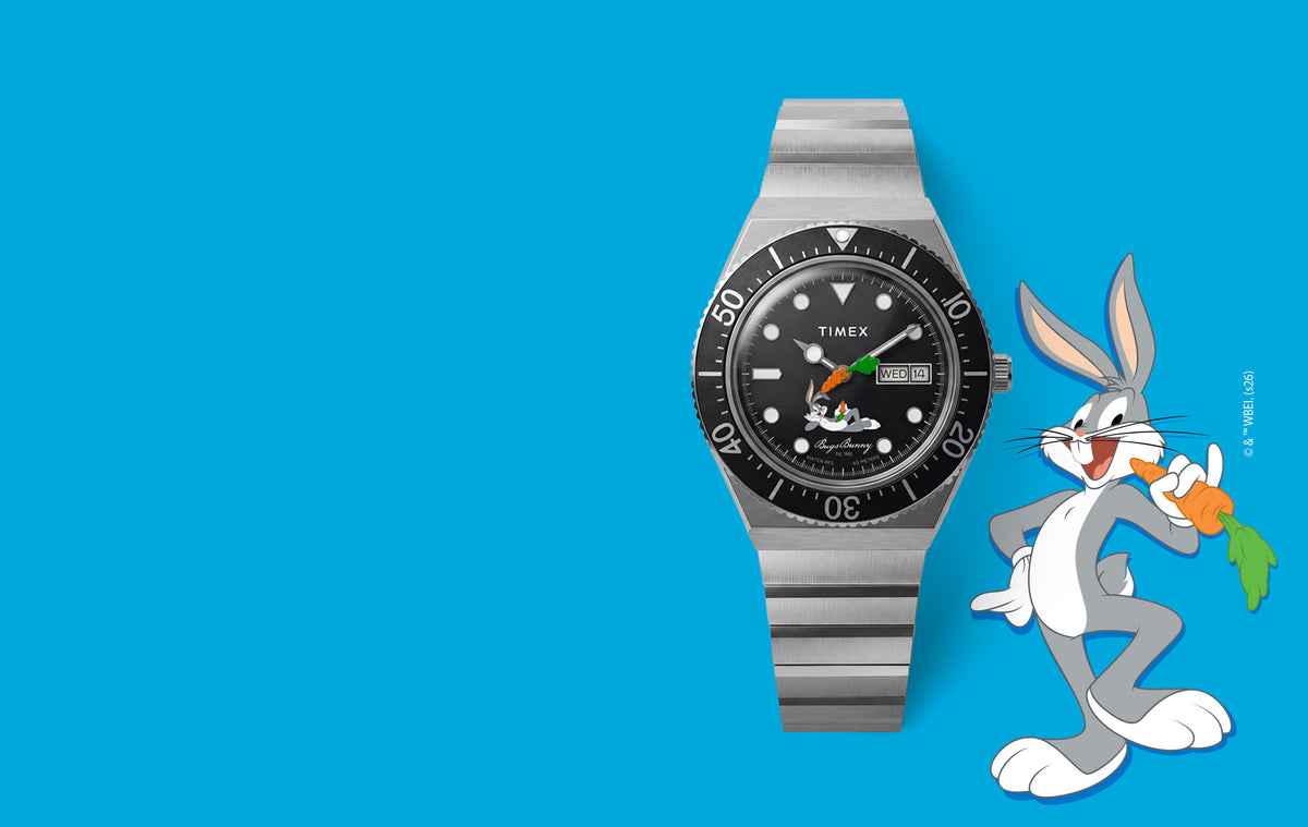 TW2Y99200 Looney Tunes Bugs Bunny x Timex M79 Automatic Signature Edition watch with a black dial, carrot-shaped hand, and stainless steel bracelet beside Bugs Bunny on a blue background.