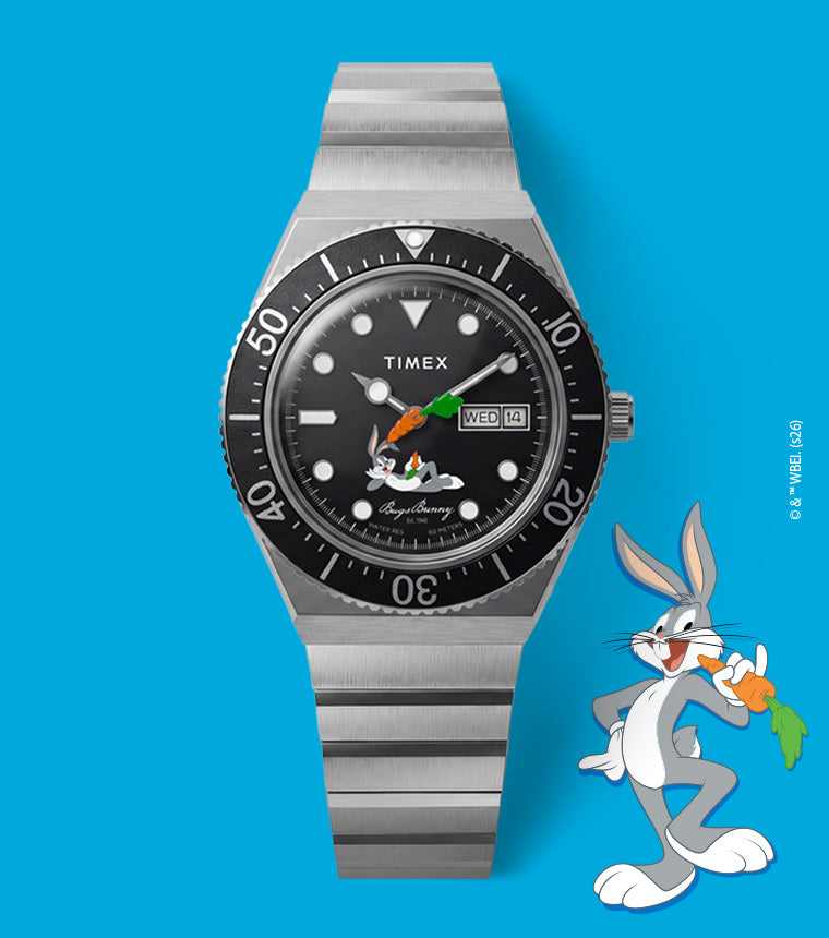 TW2Y99200 Looney Tunes Bugs Bunny x Timex M79 Automatic Signature Edition watch with a black dial, carrot-shaped hand, and stainless steel bracelet beside Bugs Bunny on a blue background.