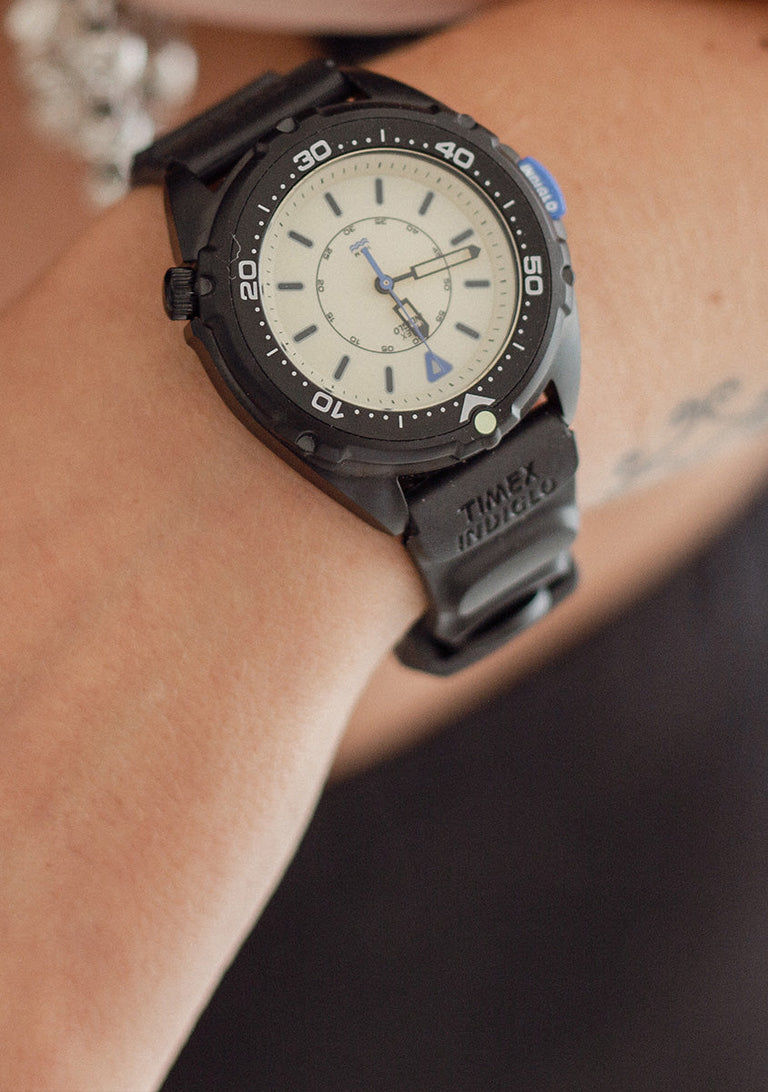 TW6A19800 Timex Intrepid x Dimepiece ‘Blackout’ Edition with a cream dial and black strap, worn on a wrist beneath a hand with silver jewelry.