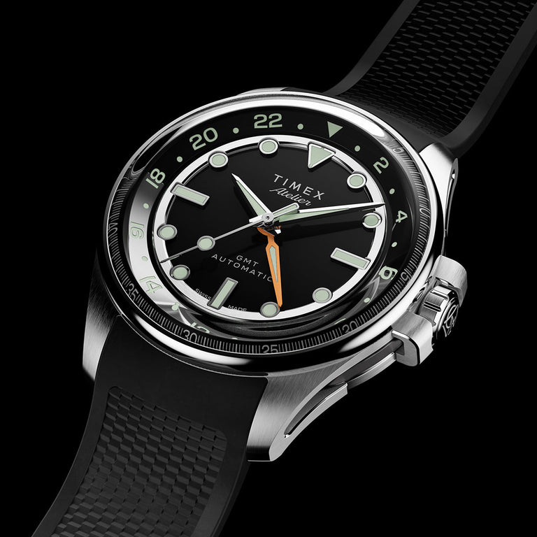 Timex Atelier GMT automatic watch with a black dial, luminous markers, orange GMT hand, and textured black rubber strap.
