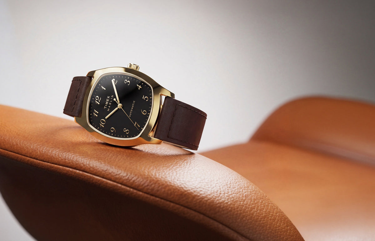 TW2Y84500 Marlin® Chatham Automatic watch with a gold-tone case, black dial and brown leather strap resting on a tan leather chair.