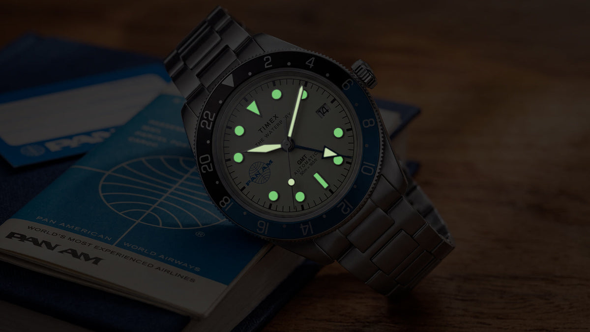 TW2Y94300 Pan Am x Timex Waterbury Automatic GMT watch glowing beside a vintage Pan Am travel book.