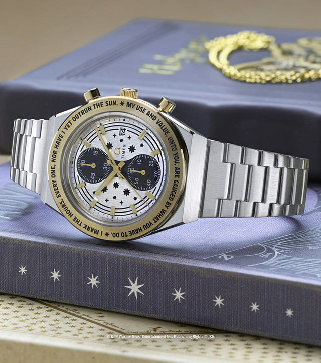 TW2Y75900 Silver-tone chronograph watch with a star-patterned dial and gold-tone bezel displayed beside a gold Time-Turner pendant on a book.
