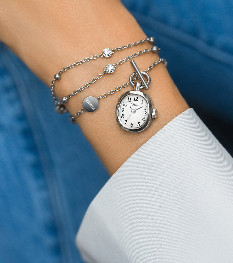 TW6A17600 Close-up of the Cavatina® Mini watch necklace worn around a wrist beside a white cuff, against blue denim.