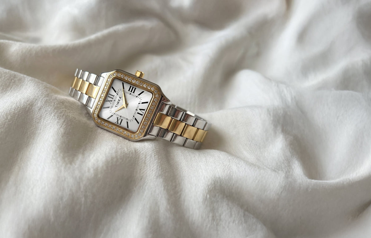 TW2Y90400 Timex Addison watch with a silver-and-gold-tone bracelet, white Roman numeral dial, and crystal-lined bezel, resting on ivory fabric.