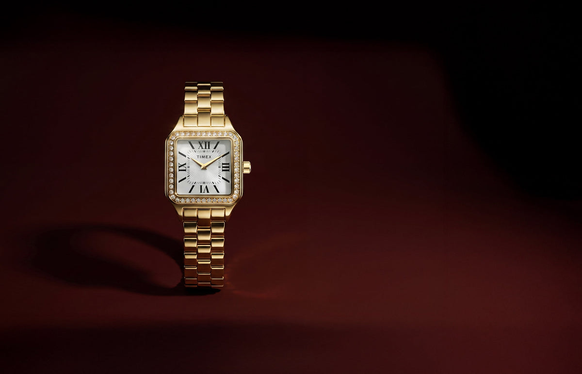 TW2Y90500 Gold-tone Timex Addison watch with a white Roman numeral dial, crystal-lined rectangular bezel, and link bracelet against a dark burgundy background.