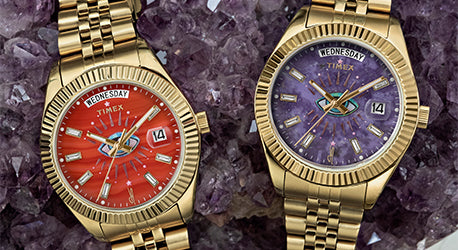 2 gold color strap watches lying across an amethyst stone. The left watch has a red agate dial and the right has an amethyst dial