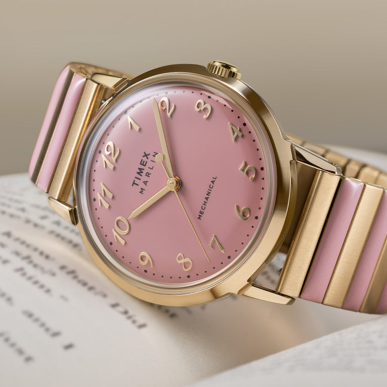 Timex women’s Marlin mechanical watch with a soft pink dial, gold-tone case, Arabic numerals, and a gold bracelet with pink enamel accents, shown in a close-up product shot.