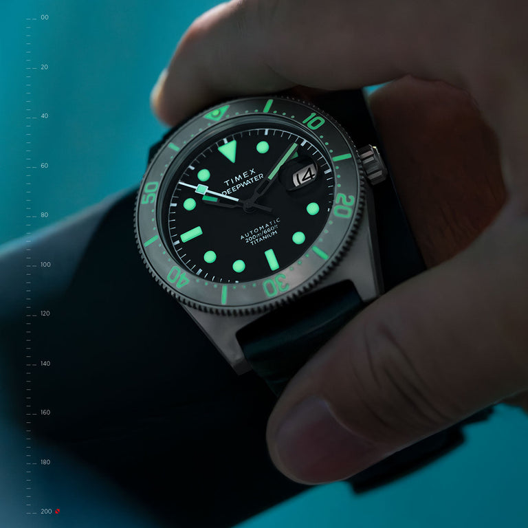 Close-up of the Timex Deepwater Automatic dive watch with luminous green markers and hands glowing underwater. The titanium case and rotating bezel are visible, with a date window at 3 o’clock. A diver’s hand adjusts the watch, emphasizing its 200-meter water resistance and rugged design.