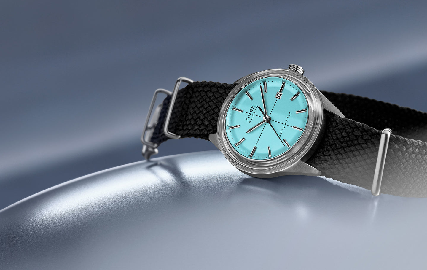 Timex Marlin Automatic watch with a vibrant turquoise dial, silver-tone case, and black textured strap, displayed on a reflective grey tone surface. 