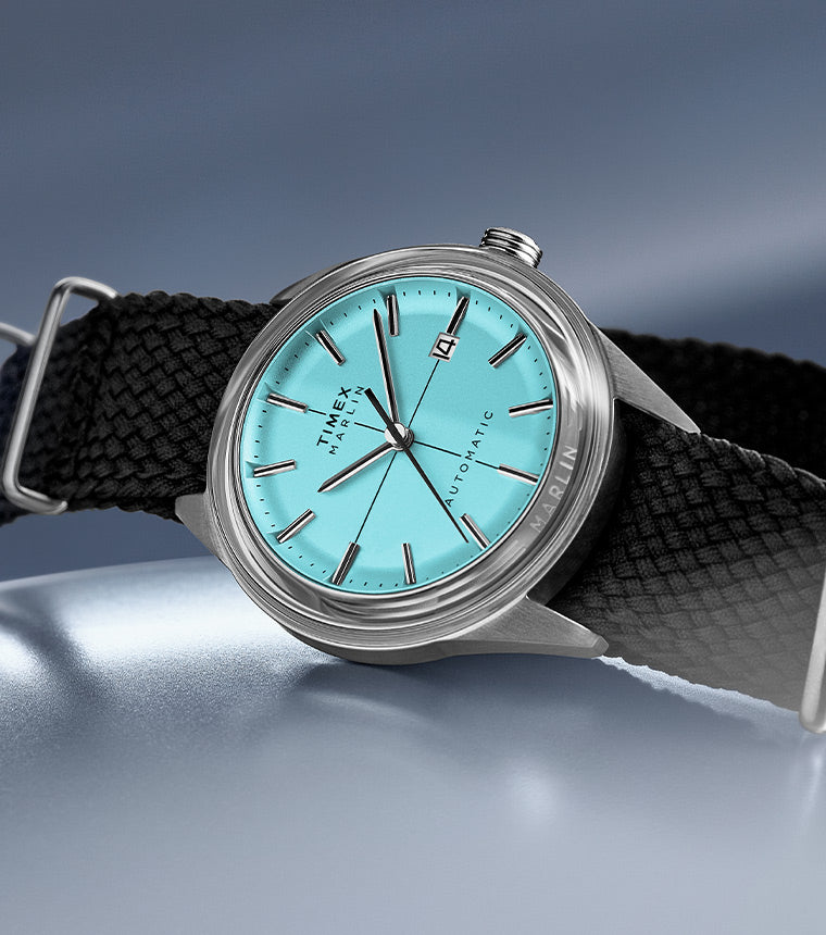 Timex Marlin Automatic watch with a vibrant turquoise dial, silver-tone case, and black textured strap, displayed on a reflective grey tone surface. 