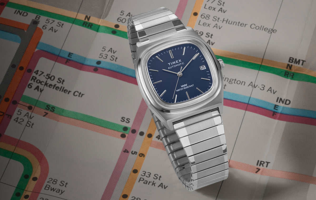 Timex Automatic 1983 E Line watch with a blue dial and stainless-steel bracelet displayed on a colorful subway map.