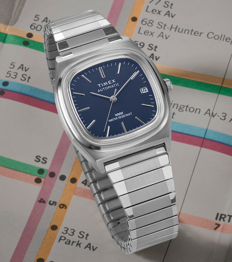 Timex Automatic 1983 E Line watch with a blue dial and stainless-steel bracelet displayed on a colorful subway map.